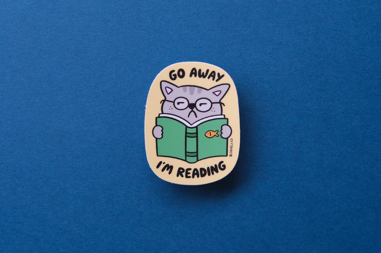 A JaneLi.Co sticker of a cat reading a book that says "Go Away I'm Reading" over a teal background.