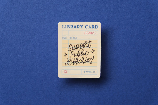 A JaneLi.Co sticker of a vintage library card that says "Support Public Libraries" over a dark blue background.