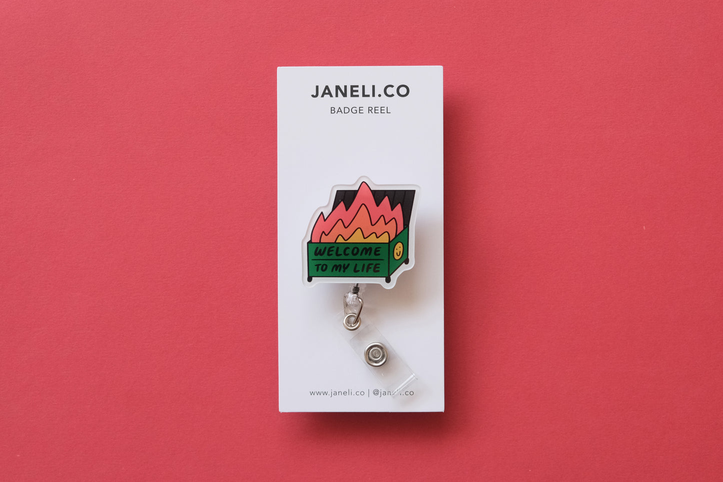 Photo of an acrylic badge reel shaped like a dumpster fire that says "Welcome to My Life" on a white backing card over a  red background.