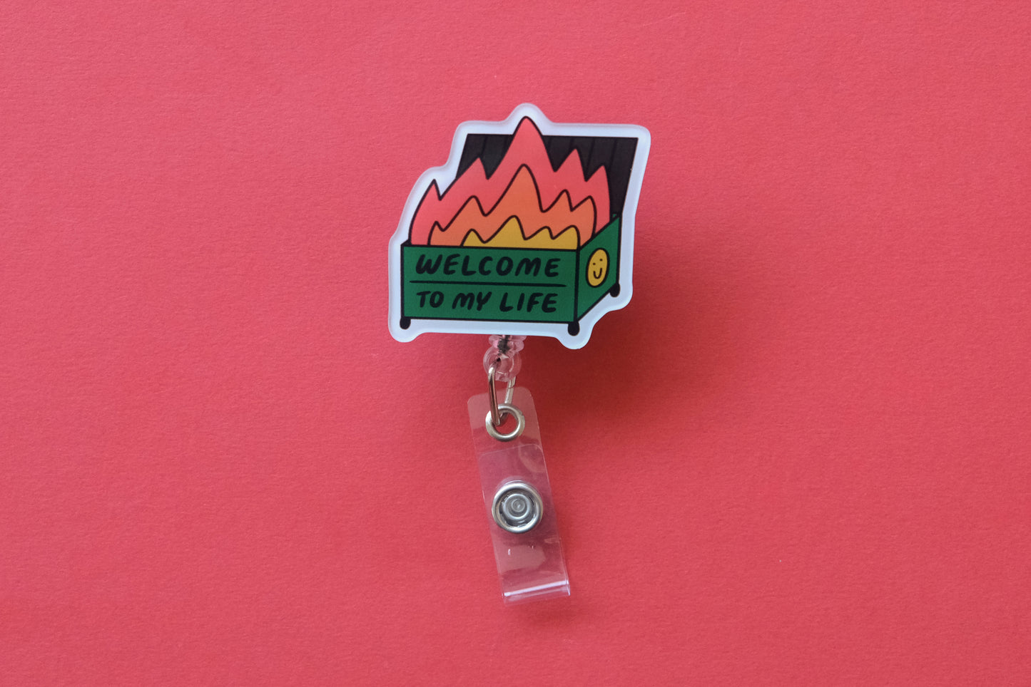 Photo of an acrylic badge reel shaped like a dumpster fire that says "Welcome to My Life" over a  red background.