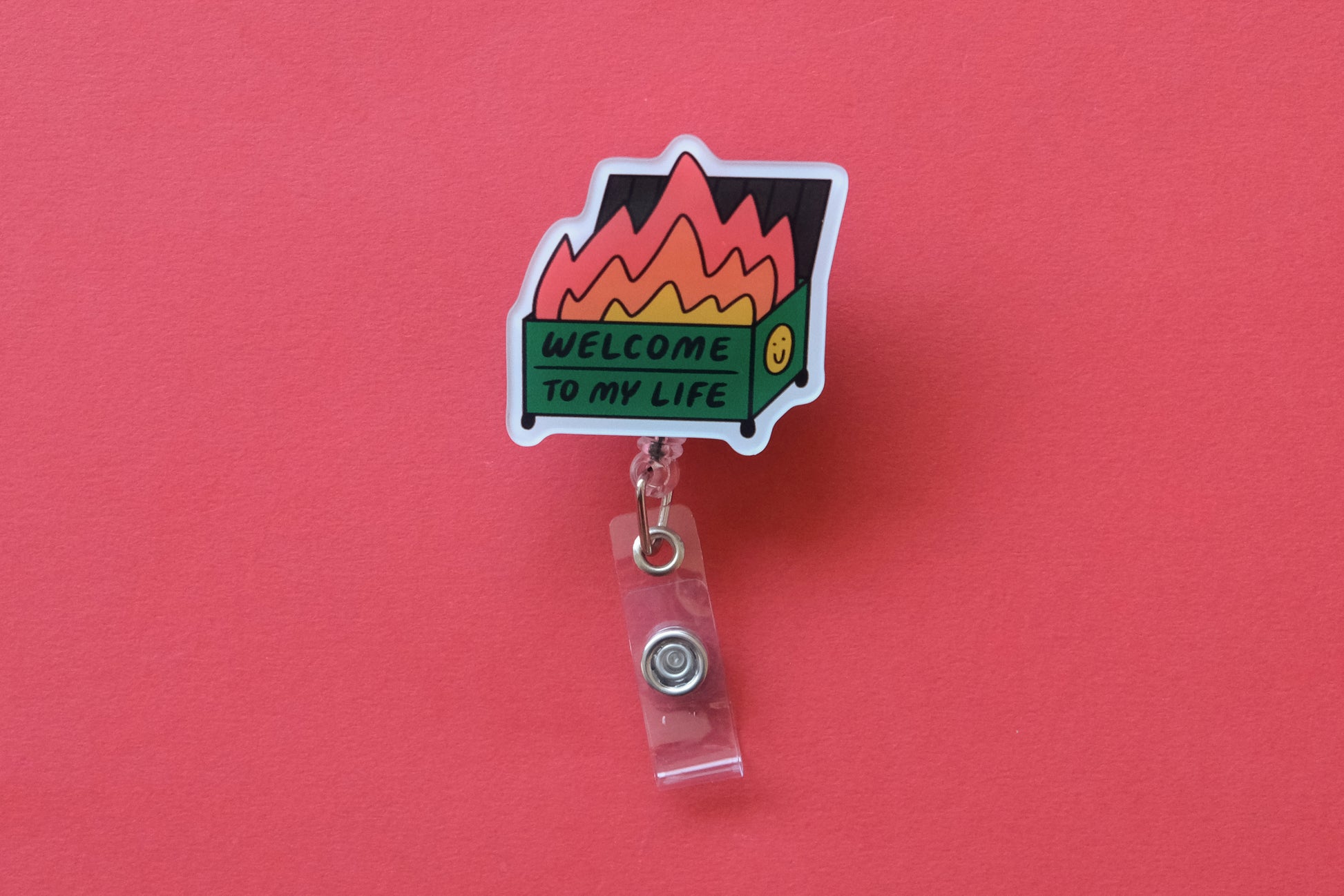 Photo of an acrylic badge reel shaped like a dumpster fire that says "Welcome to My Life" over a  red background.