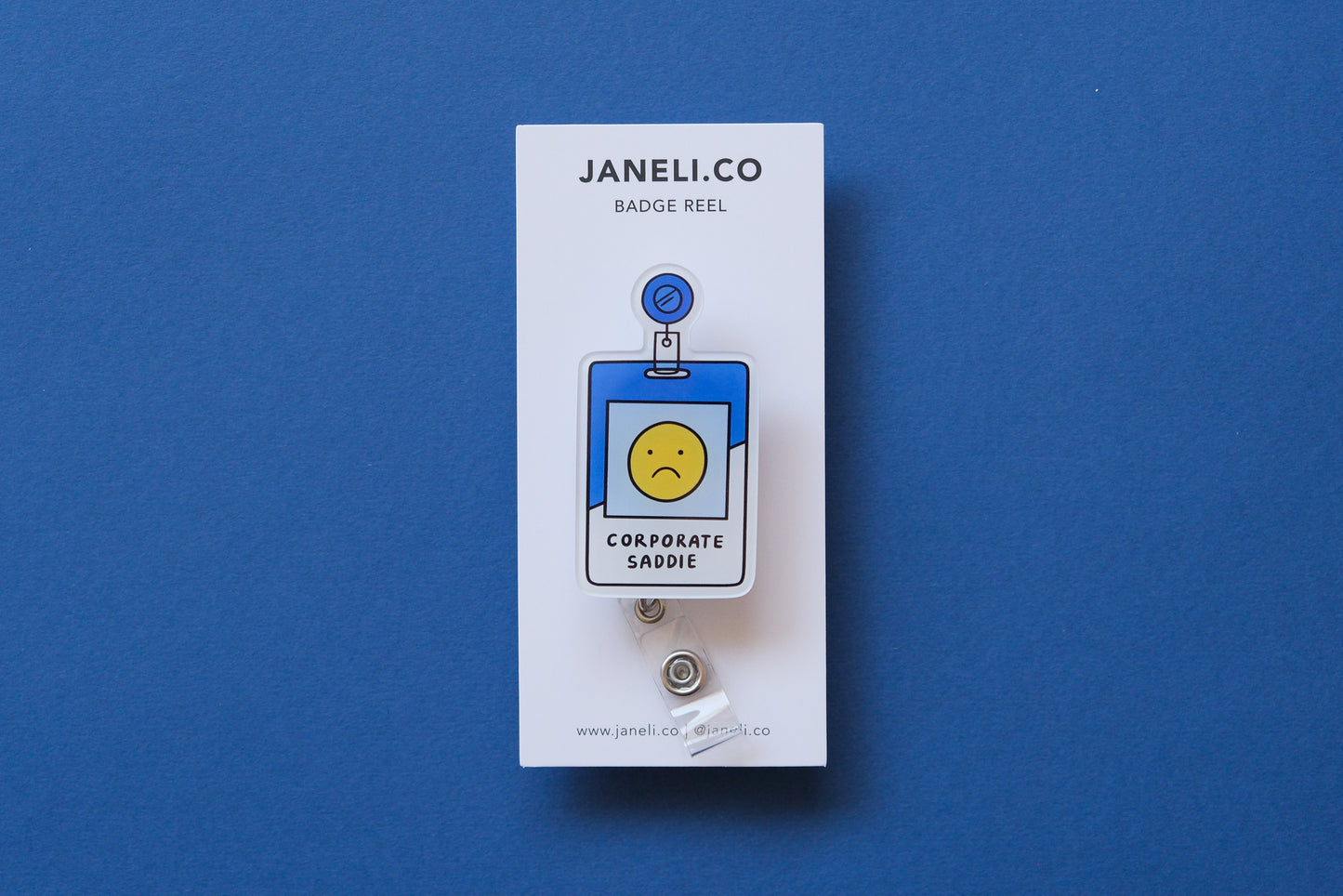 Photo of an acrylic badge reel shaped like a corporate badge that says "Corporate Saddie" on a white backing card over a dark blue background.