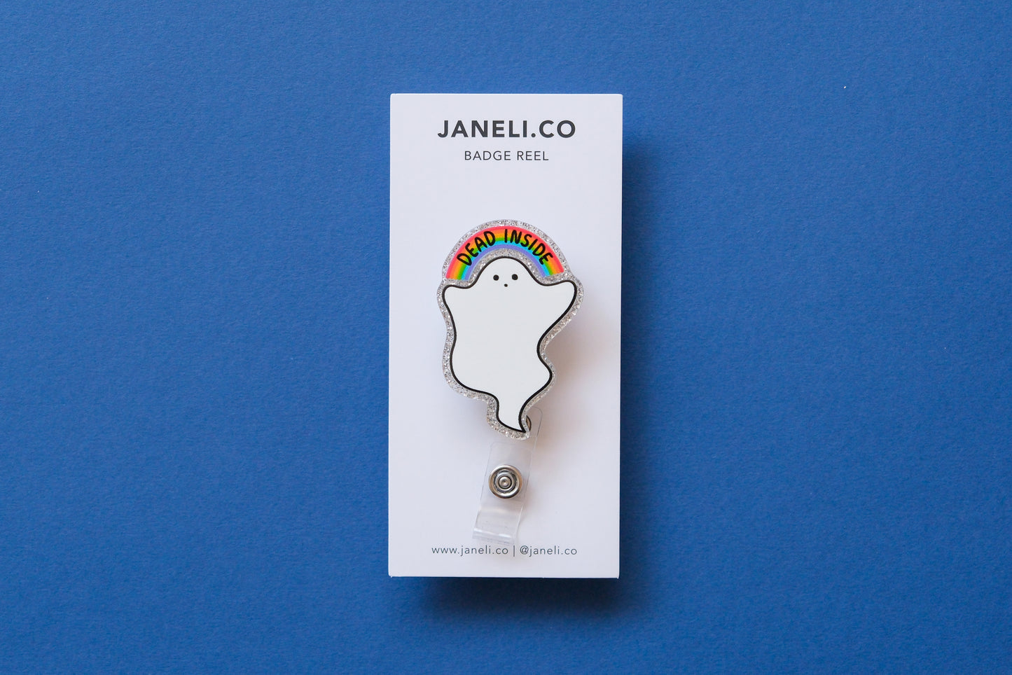 Photo of a silver glitter acrylic badge reel shaped like a ghost with a rainbow that says "Dead Inside" on a white backing card over a dark blue background.