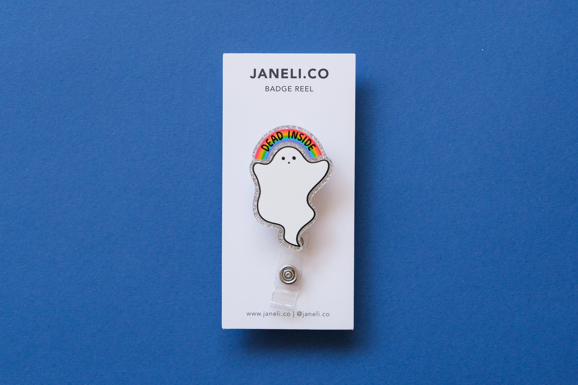 Photo of a silver glitter acrylic badge reel shaped like a ghost with a rainbow that says "Dead Inside" on a white backing card over a dark blue background.