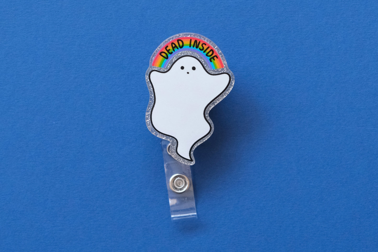Photo of a silver glitter acrylic badge reel shaped like a ghost with a rainbow that says "Dead Inside" over a dark blue background.