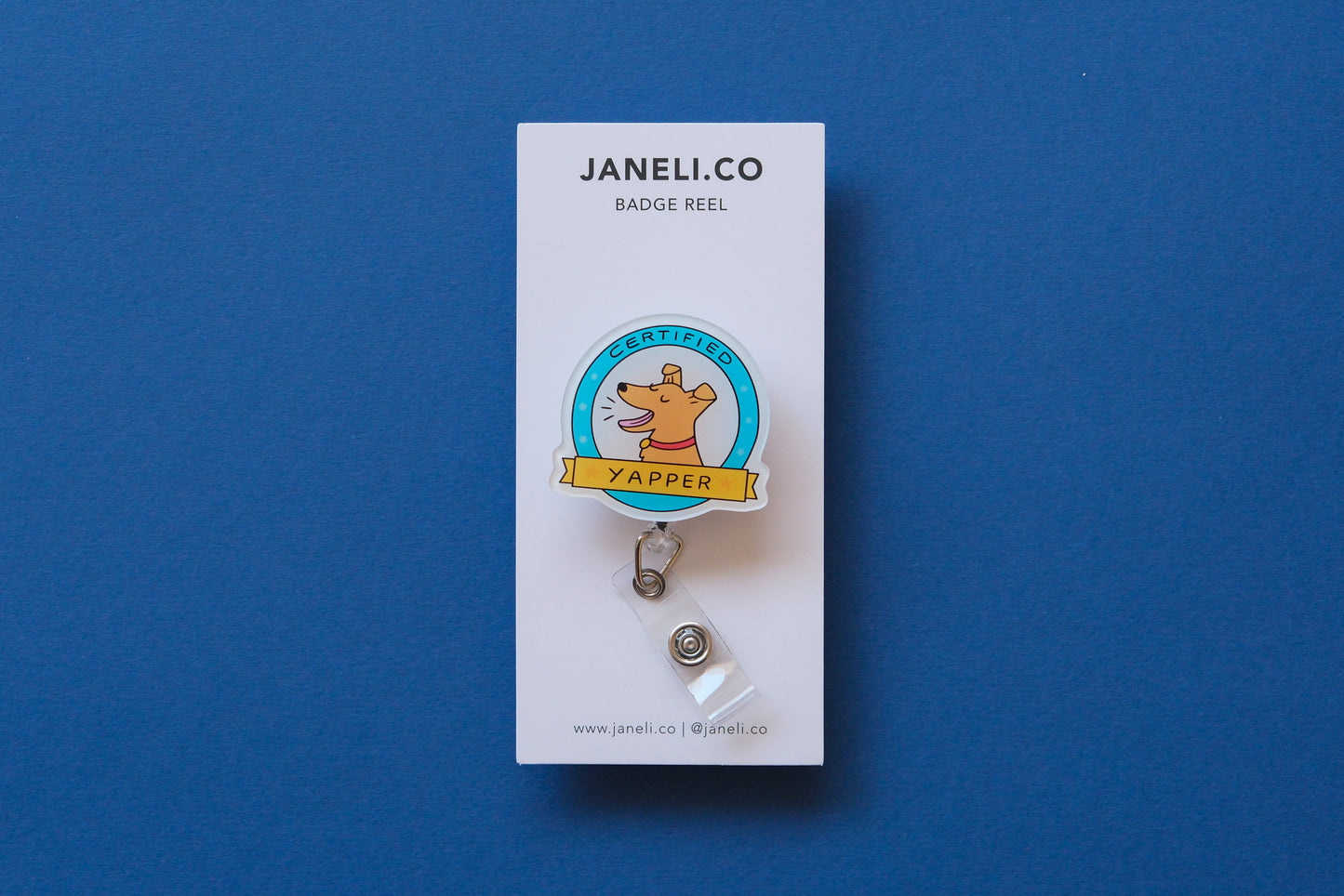Photo of an acrylic badge reel shaped like a dog badge that says "Certified Yapper" on a white backing card over a blue background.