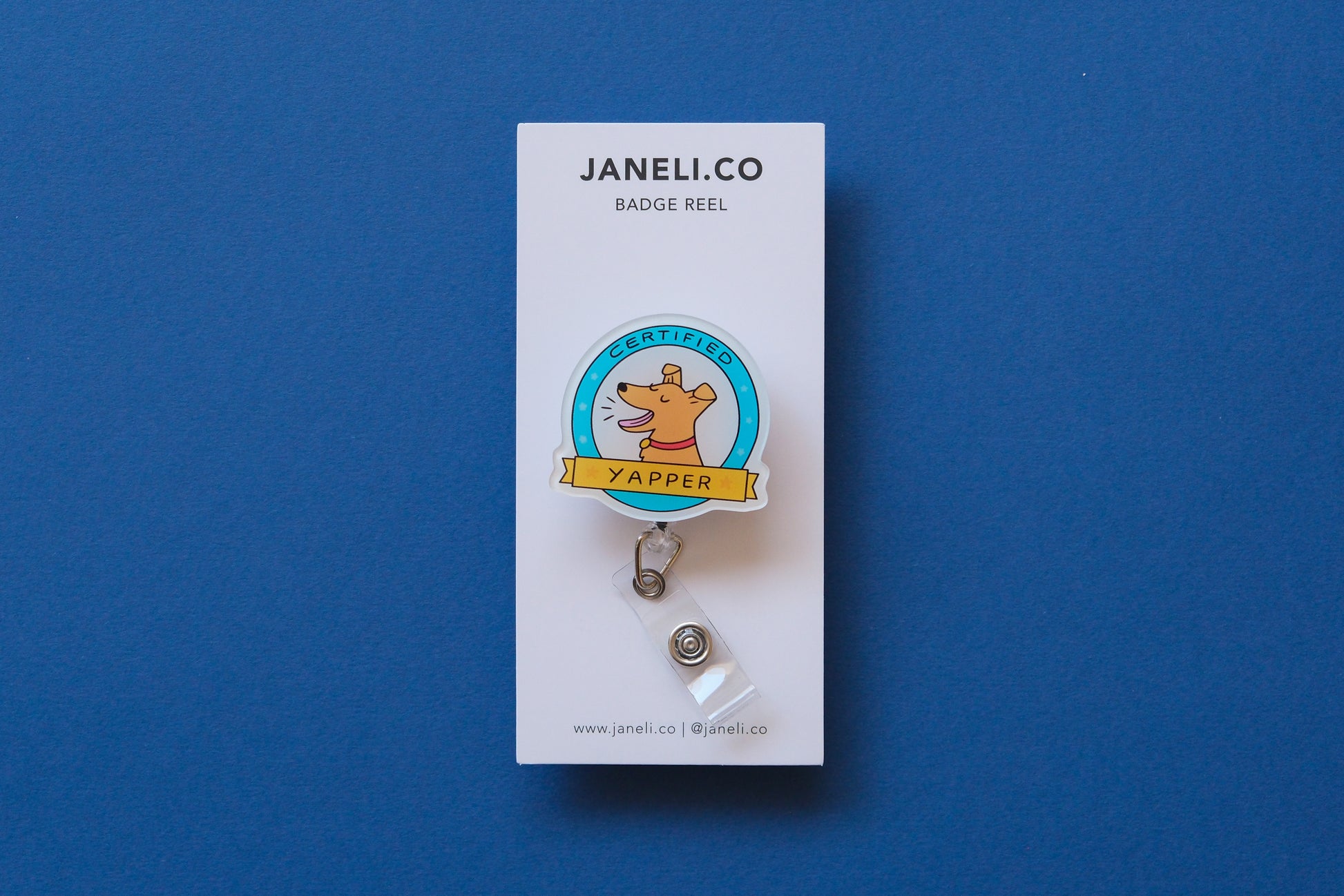 Photo of an acrylic badge reel shaped like a dog badge that says "Certified Yapper" on a white backing card over a blue background.