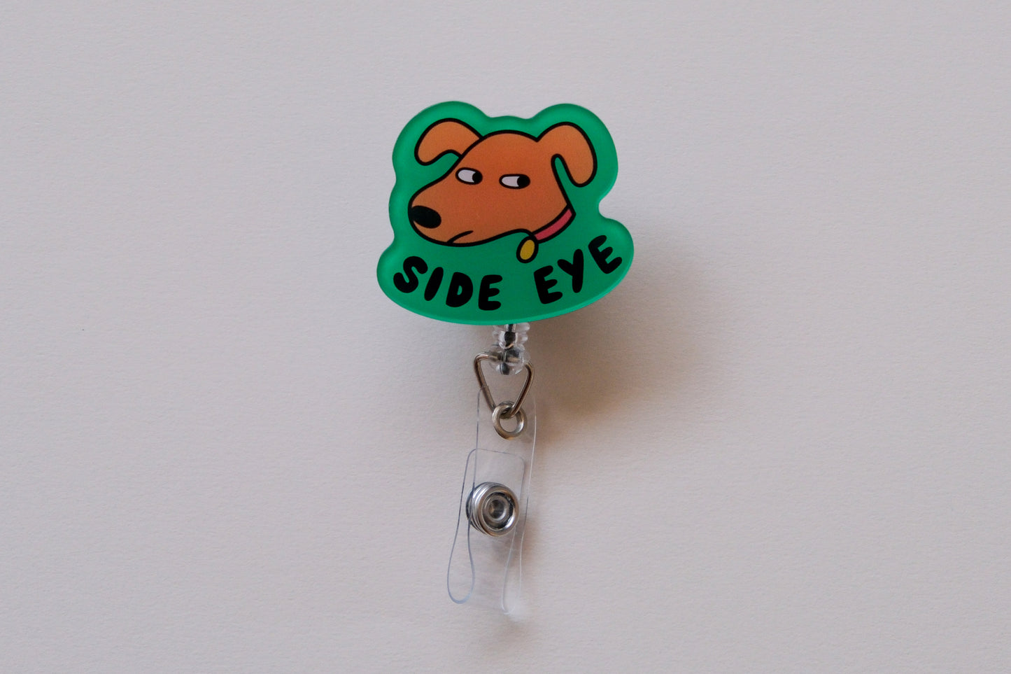 Photo of an acrylic badge reel shaped like a dog that says "Side Eye" over a cream background.