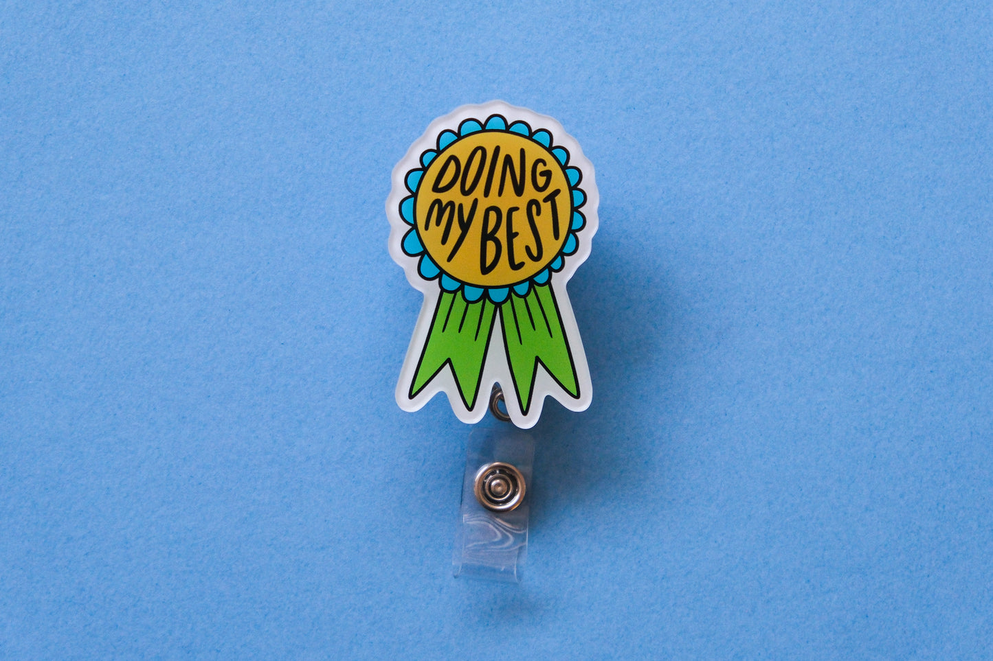Photo of an acrylic badge reel shaped like an award ribbon that says "Doing My Best" over a light blue background.