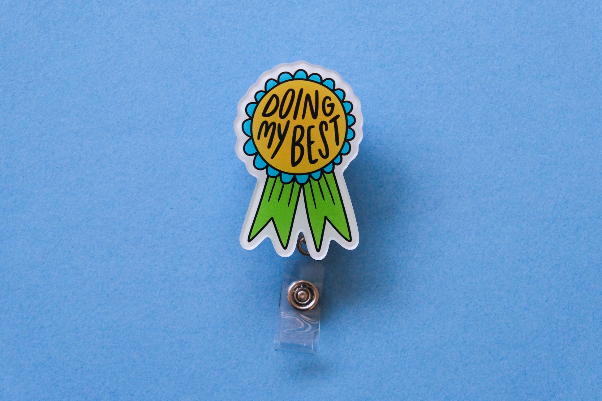 Photo of an acrylic badge reel shaped like an award ribbon that says "Doing My Best" over a light blue background.