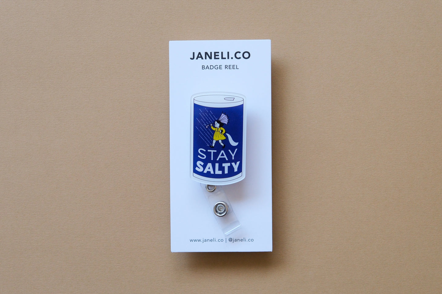 Photo of an acrylic badge reel shaped like salt can that says "Stay Salty" on a white backing card over a tan background.