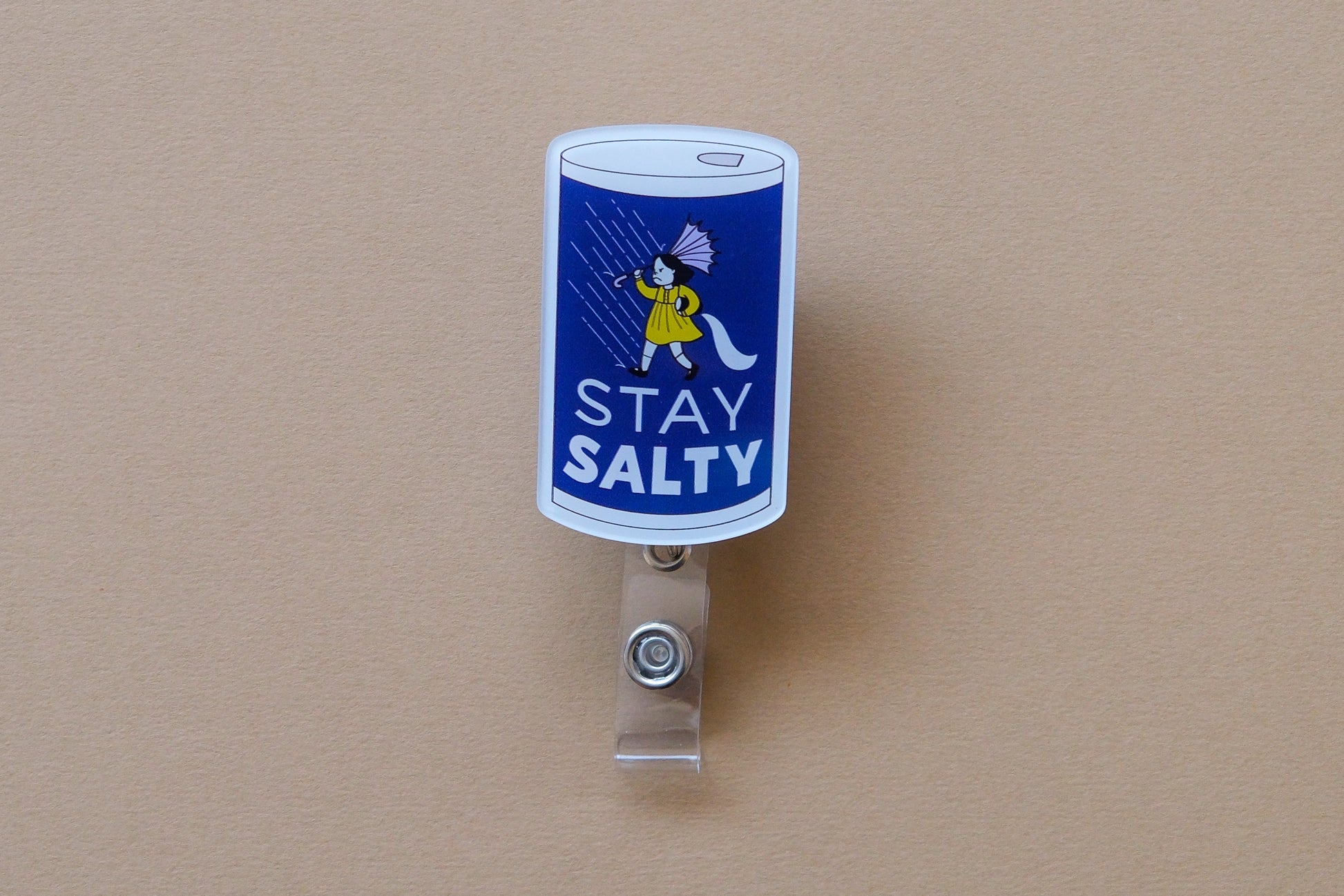 Photo of an acrylic badge reel shaped like salt can that says "Stay Salty" over a tan background.
