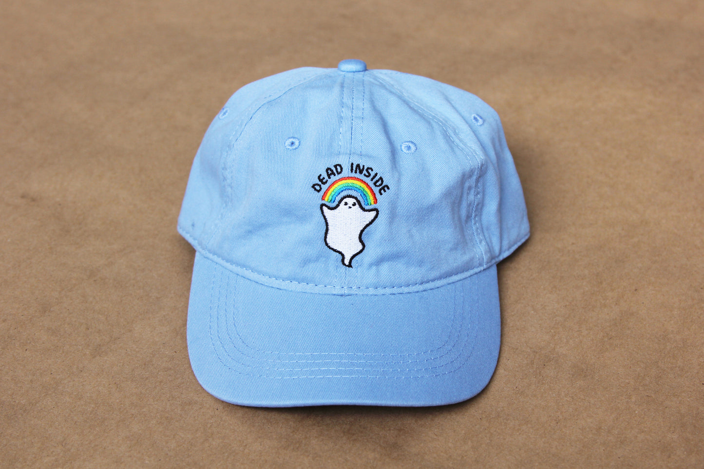 A photo of a light blue colored dad hat with an embroidered rainbow and ghost that says "Dead Inside" over a tan background.