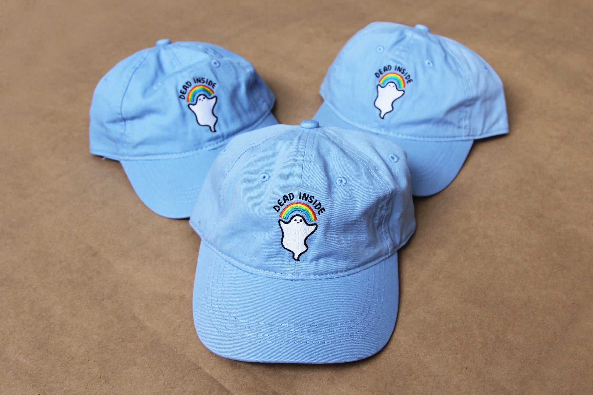 A photo of three light blue colored dad hats with an embroidered rainbow and ghost that says "Dead Inside" over a tan background.