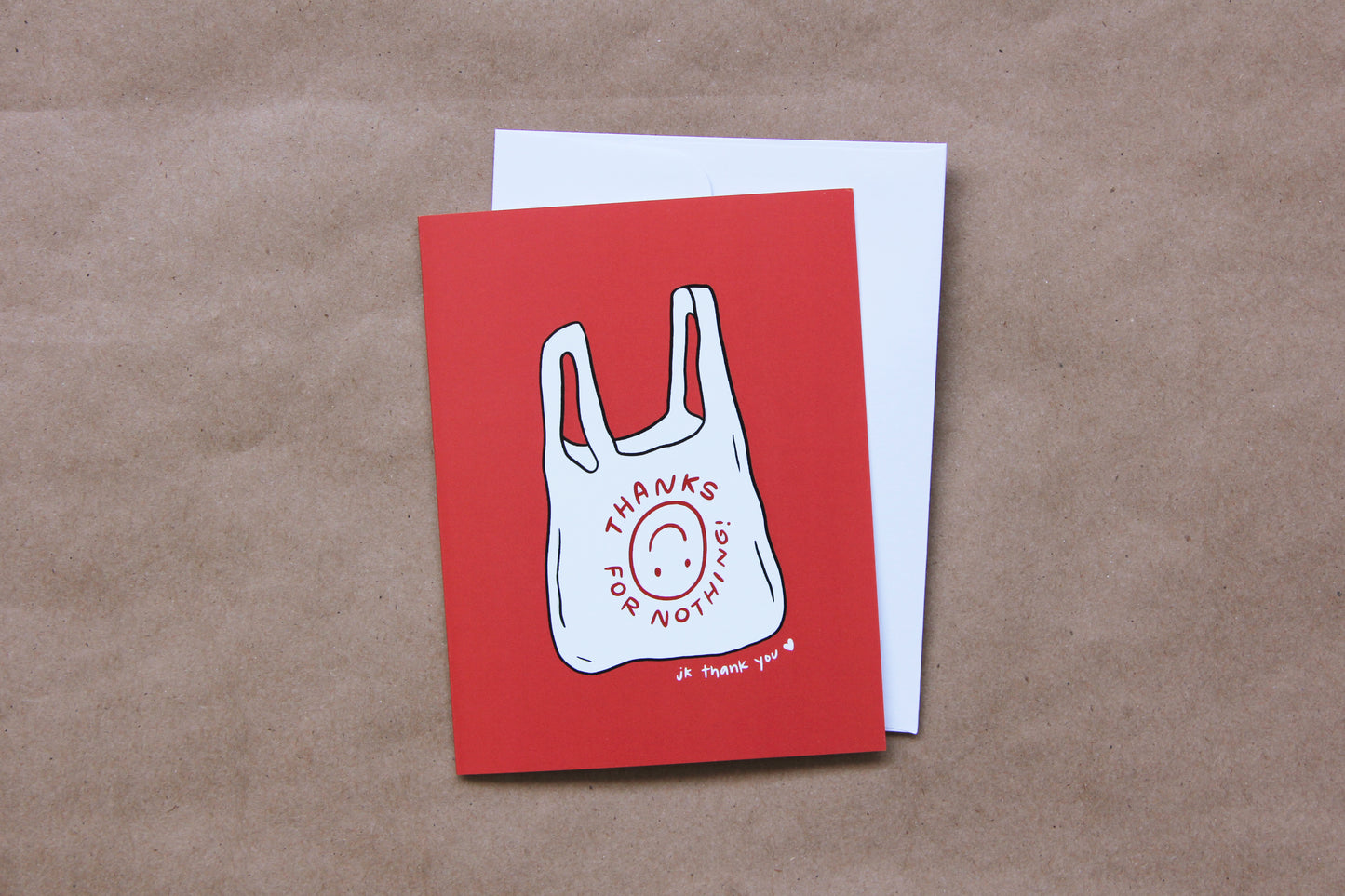 A photo of a red greeting card with a takeout bag that says "Thanks For Nothing jk thank you <3" and a white envelope on a tan background.
