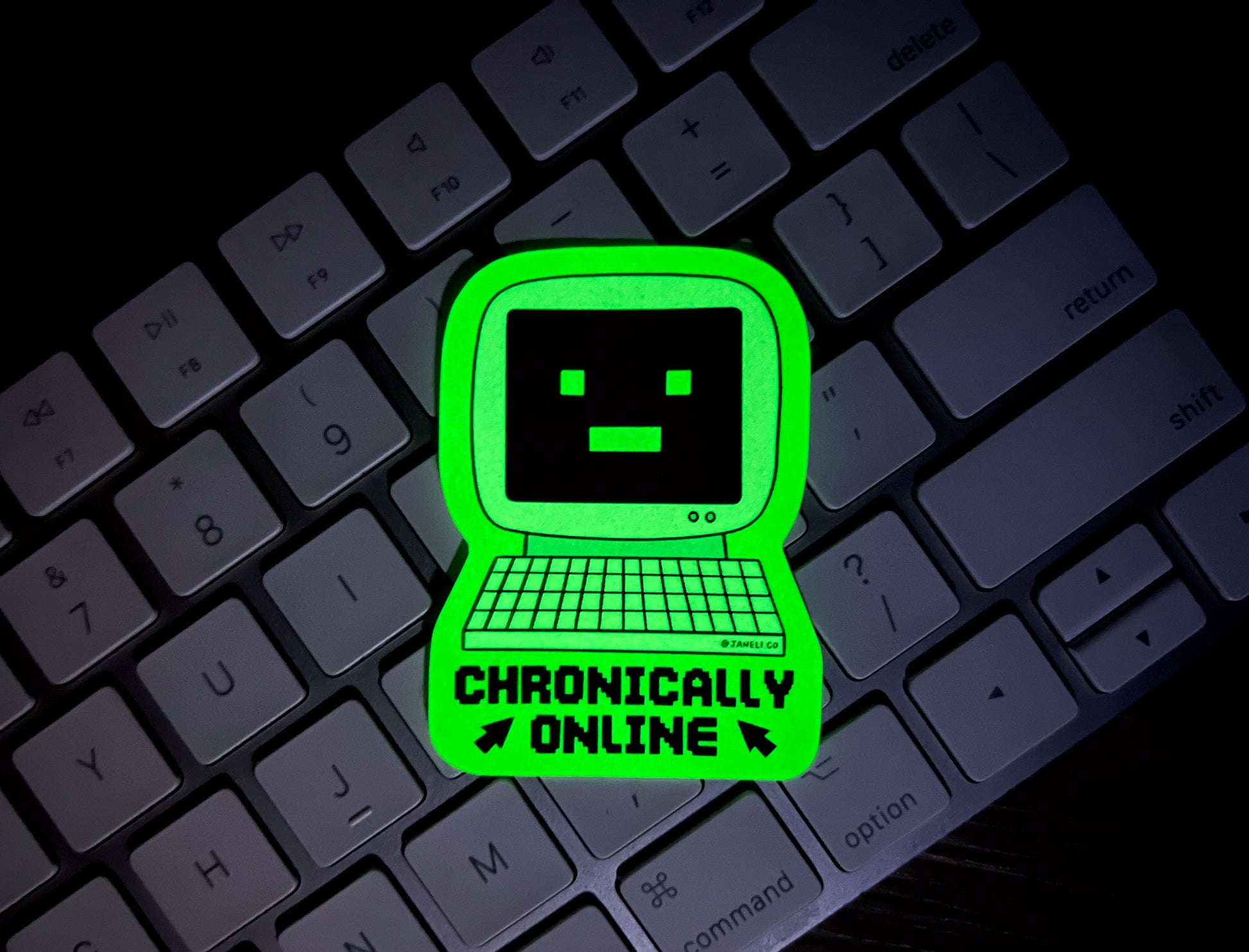 A glow in the dark sticker of a retro computer with a -_- face that says "Chronically Online" over a keyboard in the dark.