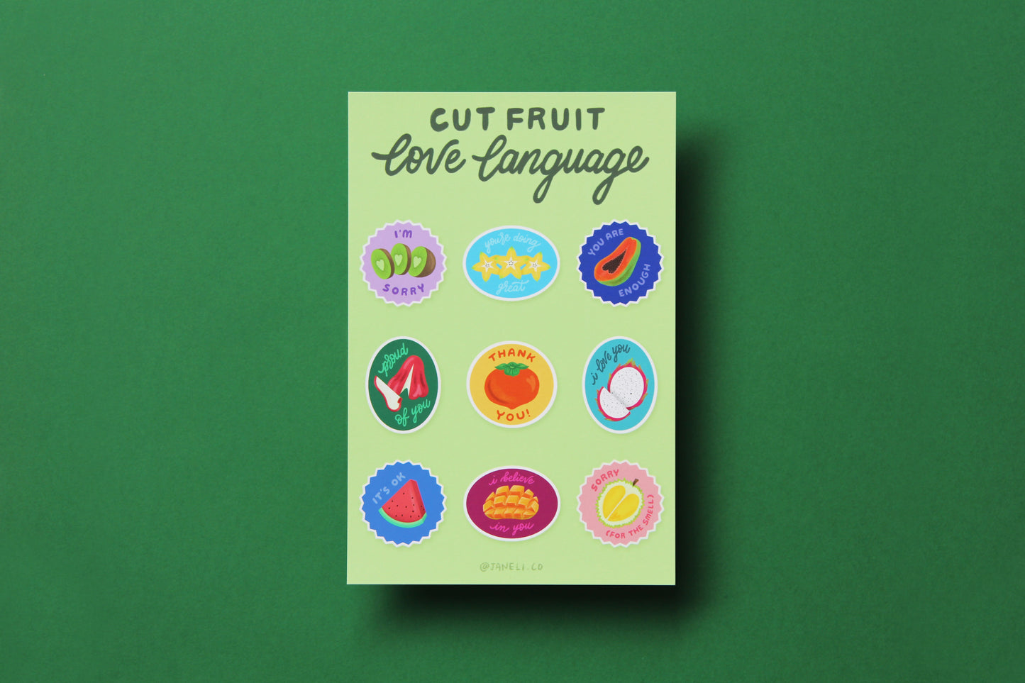 A JaneLi.Co mini print/postcard that says "Cut Fruit Love Language" with 9 different fruit stickers with different messages like "I love you", "I'm sorry", and "I'm proud of you" over a green background.