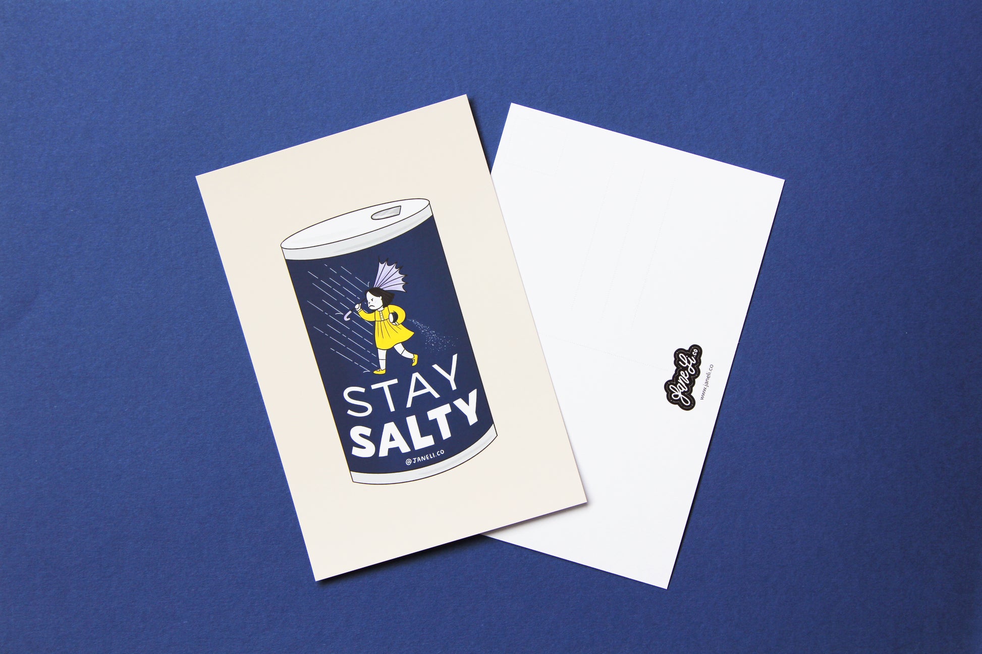 A JaneLi.Co mini print/postcard that shows a grumpy Morton salt girl spilling a can of salt in the rain and a back postcard side of the same print over a navy background.