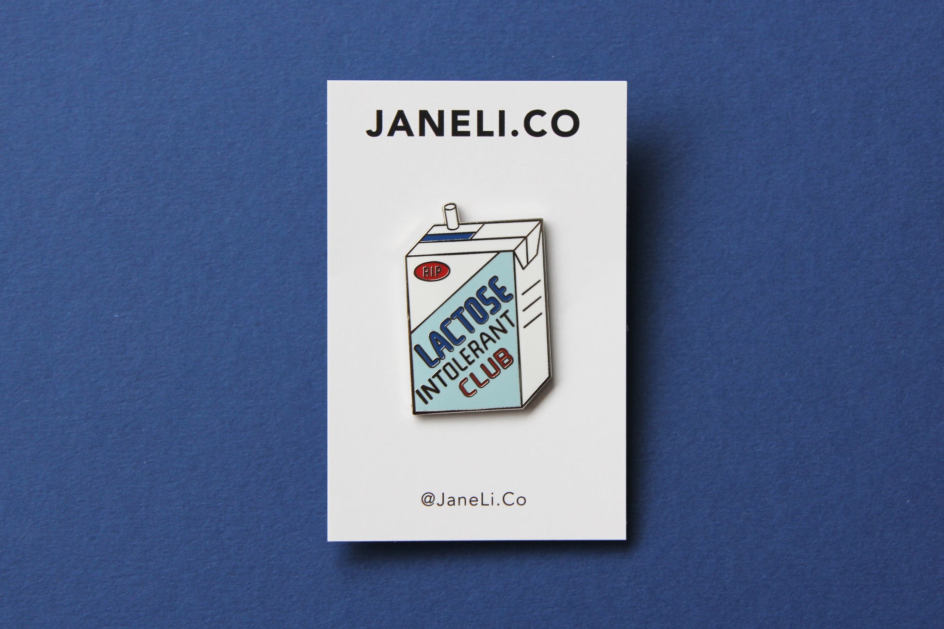 An enamel pin showing a soymilk carton that says" Lactose Intolerant Club" on a white JaneLi.Co backing card over a blue background.