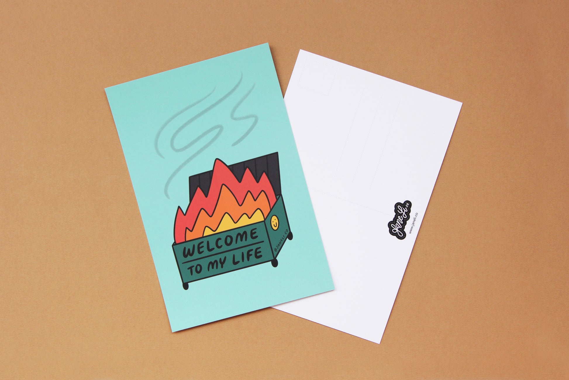 A JaneLi.Co mini print/postcard of a dumpster fire that says "Welcome to My Life" and a back postcard side of the same print over a tan background.