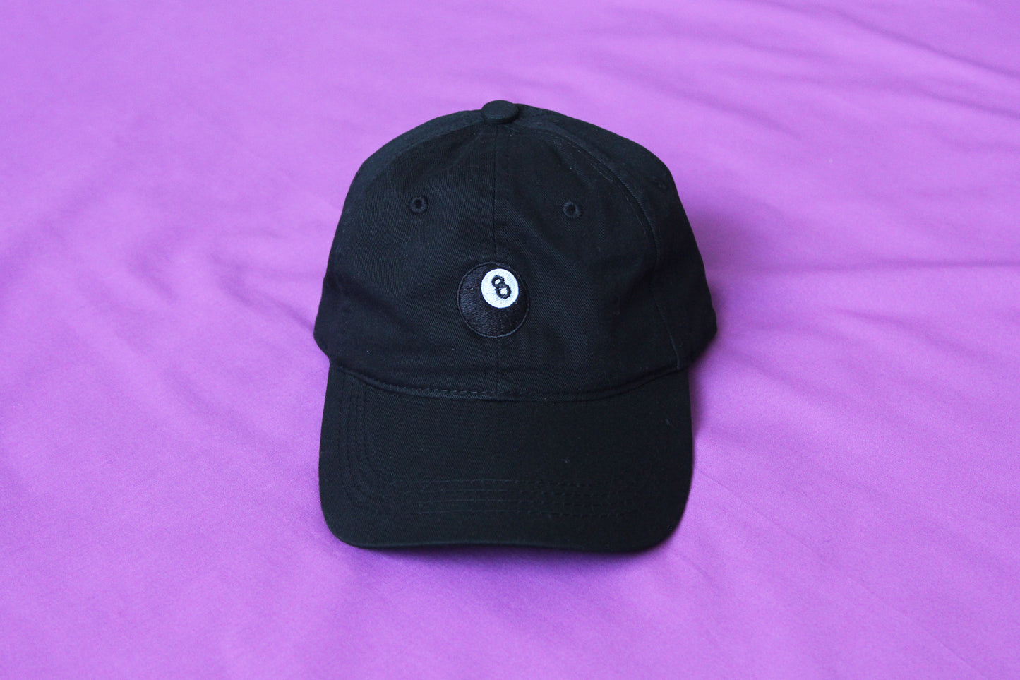 A photo of a black dad hat with an embroidered magic 8 ball over a purple background.