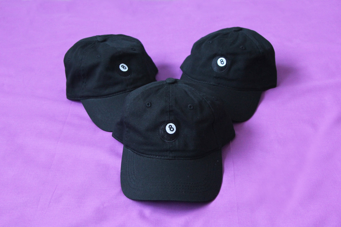 A photo of 3 black dad hats with an embroidered magic 8 ball over a purple background.