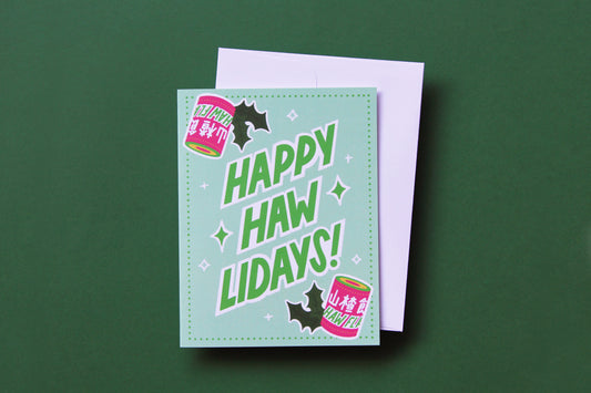 A photo of a greeting card with haw flakes and lettering that says "Happy Haw-lidays!" and a white envelope on a green background.