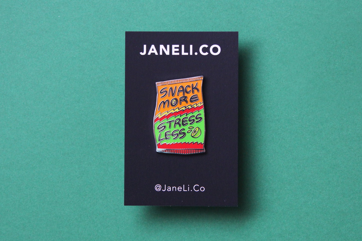 An enamel pin showing a chip bag that says "Snack More Stress Less" on a black JaneLi.Co backing card over a green background.