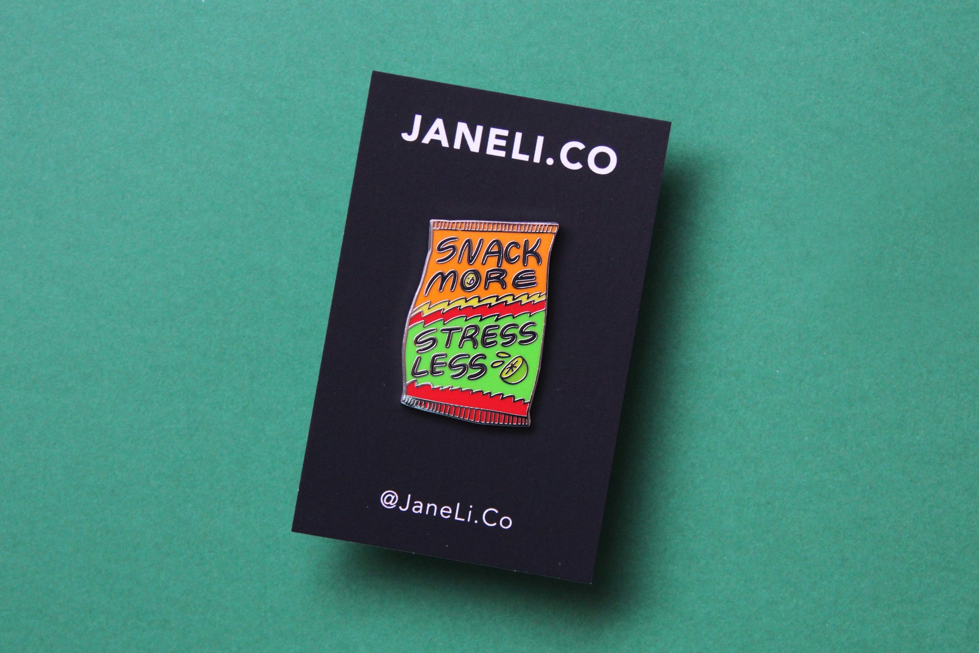 An enamel pin showing a chip bag that says "Snack More Stress Less" on a black JaneLi.Co backing card over a green background.