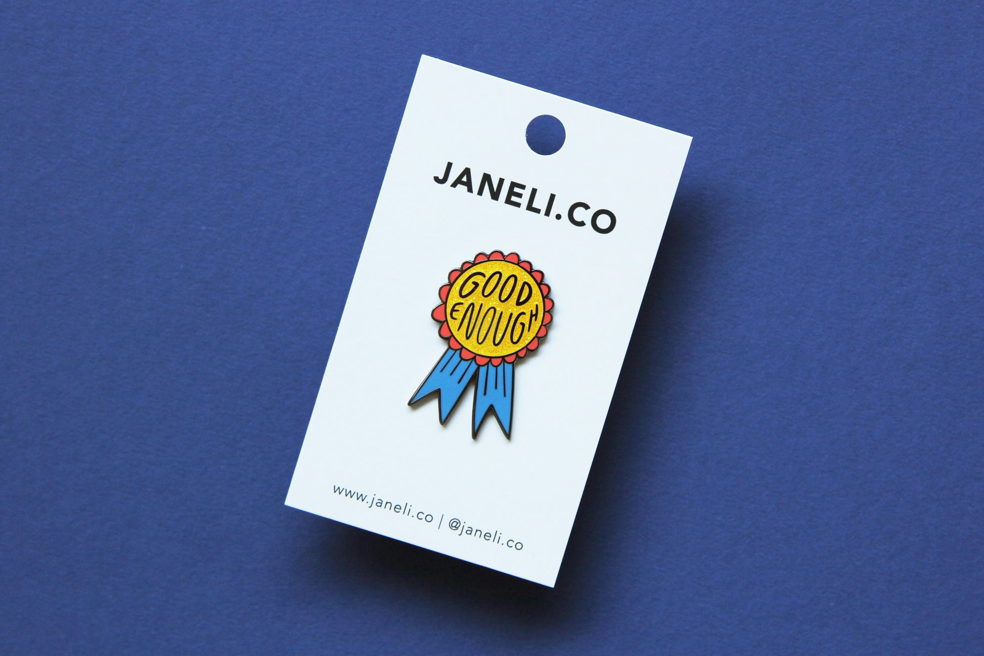 An enamel pin showing a glittery red and blue award ribbon that says "Good Enough" on a white JaneLi.Co backing card over a blue background.