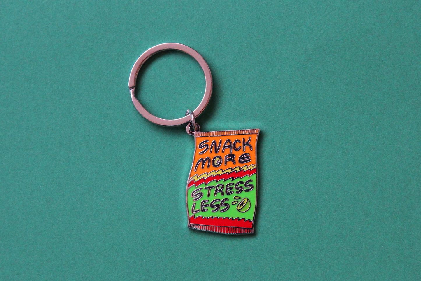 An enamel keychain showing a chip bag that says "Snack More Stress Less" over a green background.