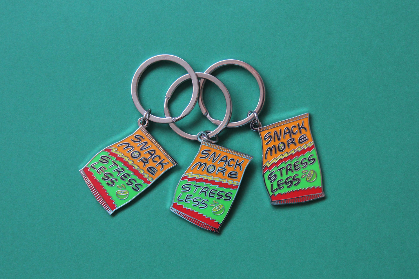 3 enamel keychains showing a chip bag that says "Snack More Stress Less" over a green background.