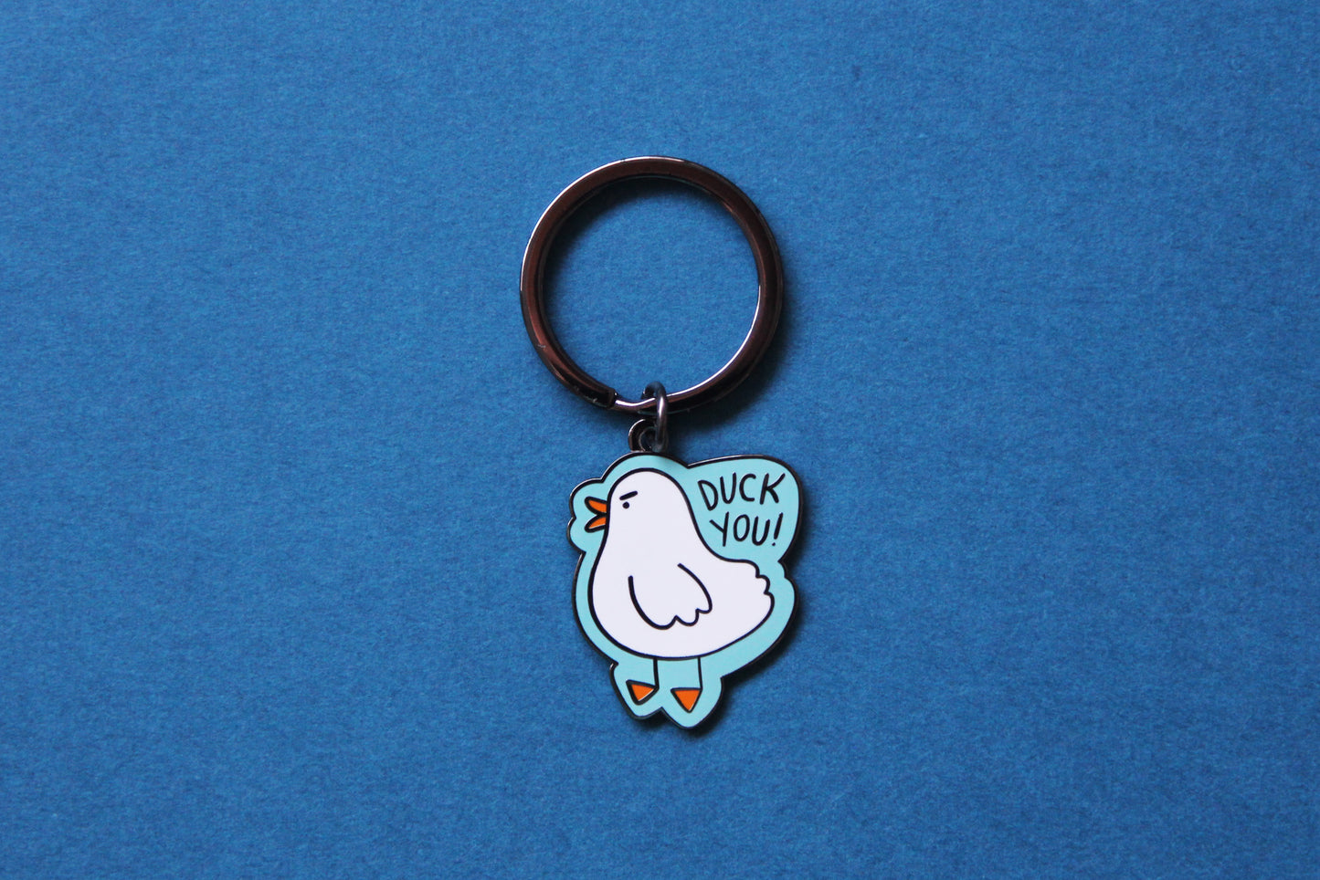 An enamel keychain showing an angry white duck saying "Duck you!" over a teal background.