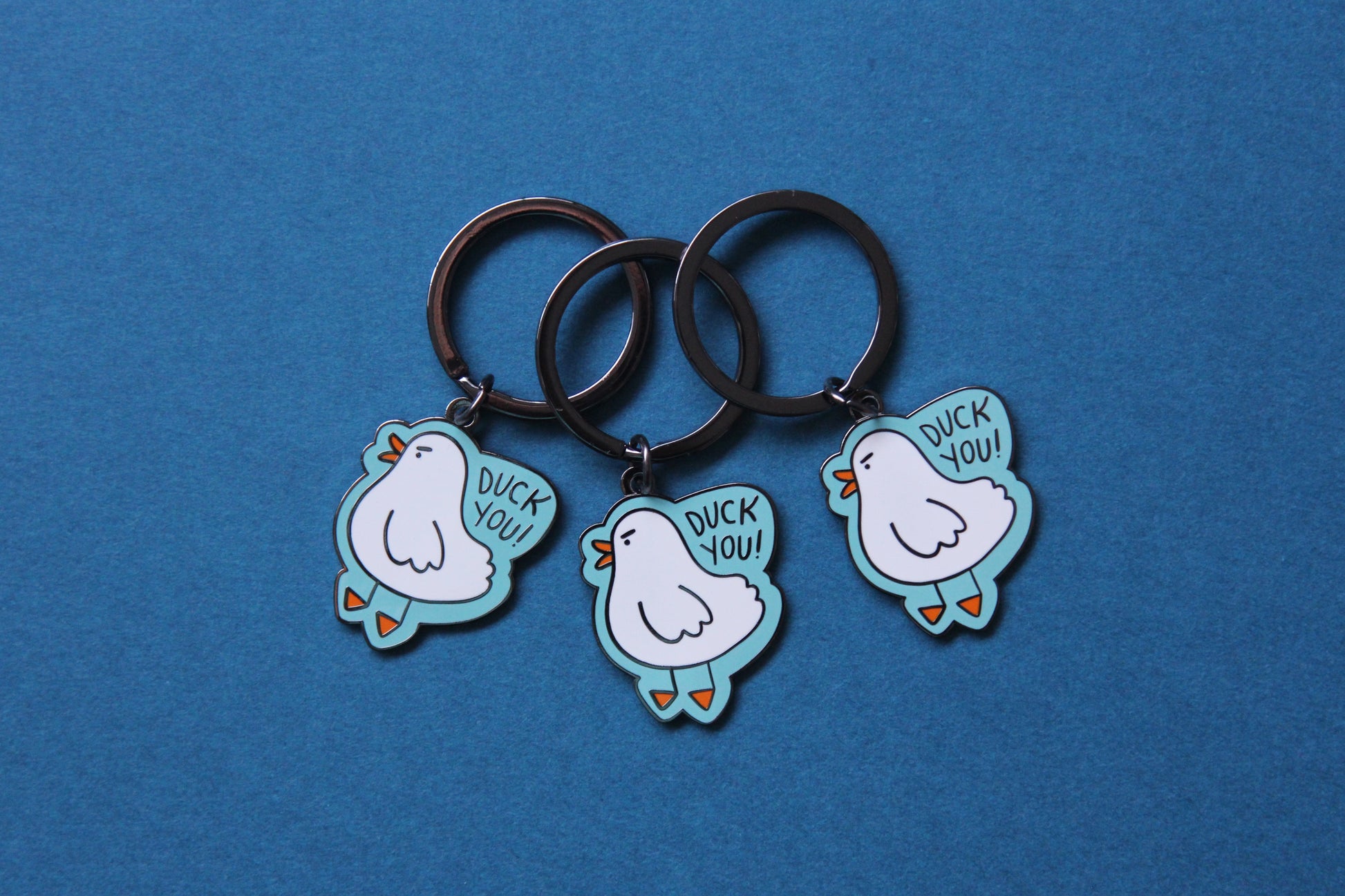 3 enamel keychains showing an angry white duck saying "Duck you!" over a teal background.