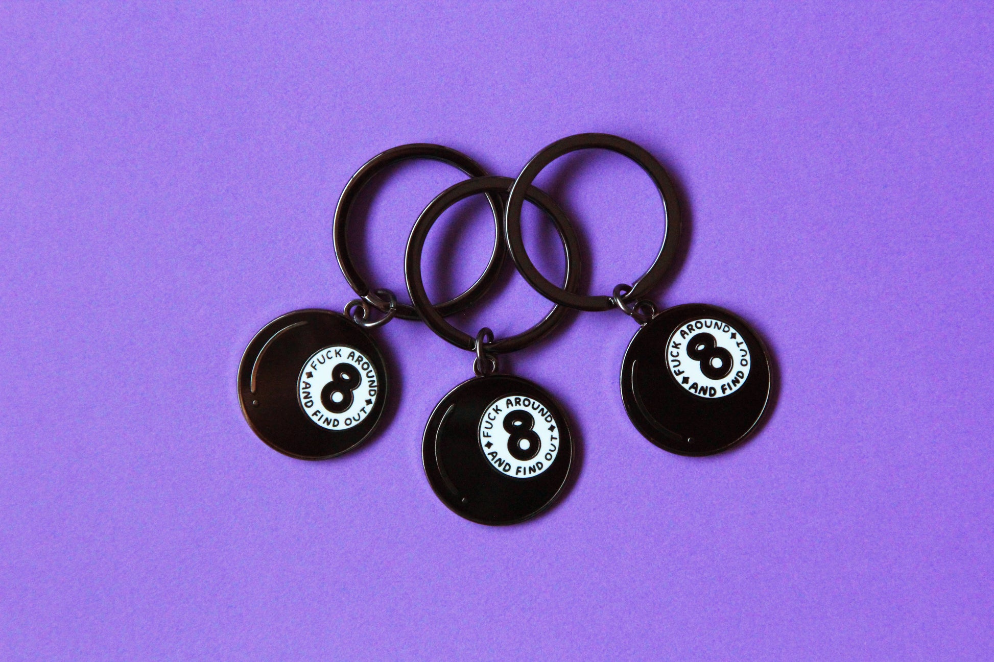 3 enamel keychains showing a magic 8 ball that says "Fuck around and find out" over a purple background.