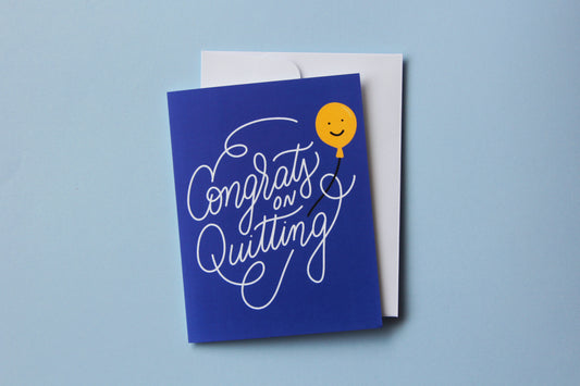 A photo of a blue greeting card with lettering that says "Congrats on Quitting" with a yellow smiley face balloon and a white envelope on a blue background.