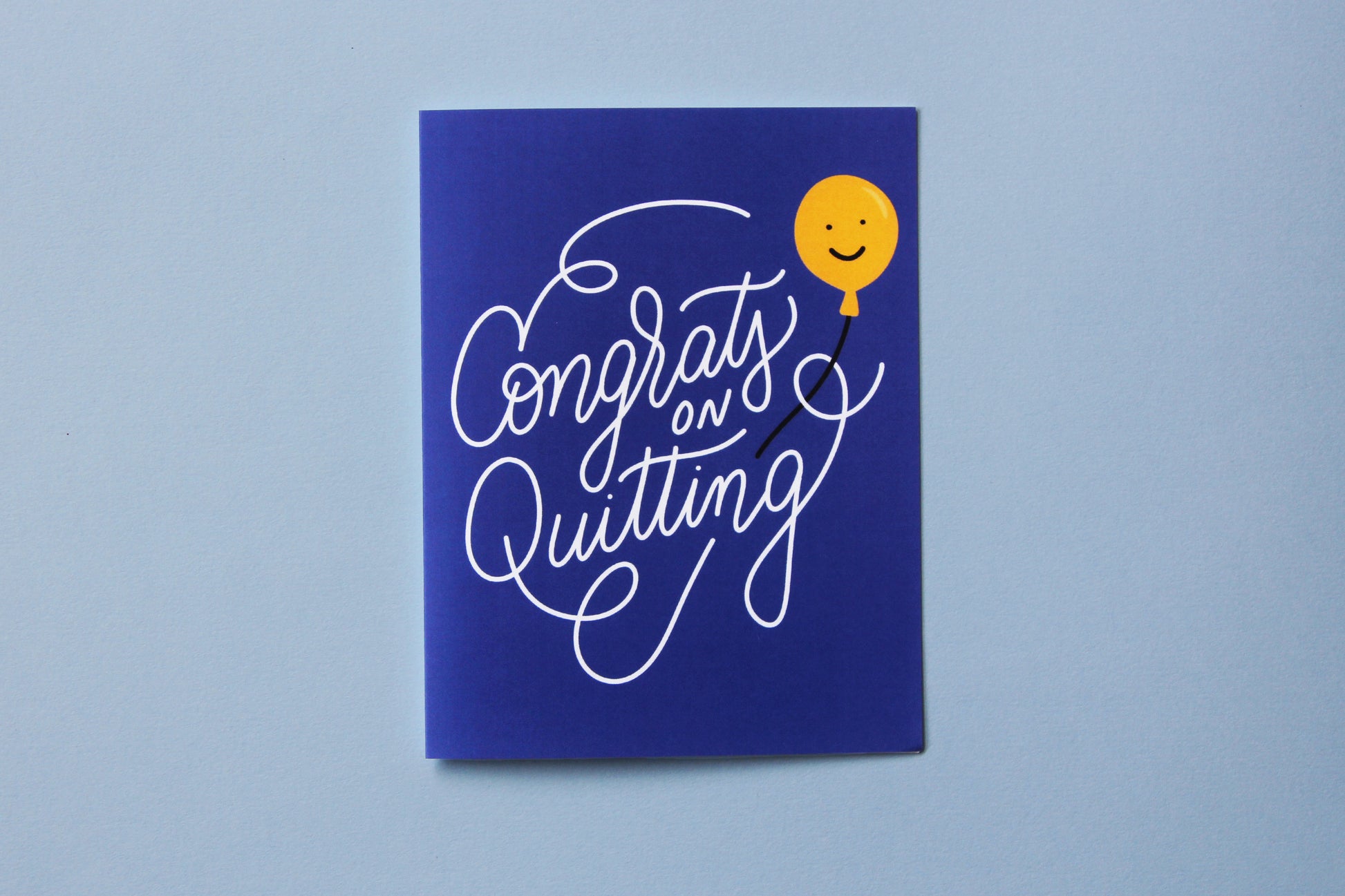 A photo of a blue greeting card with lettering that says "Congrats on Quitting" with a yellow smiley face balloon on a blue background.
