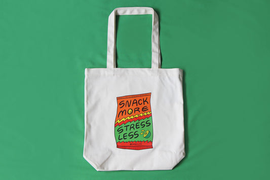 A photo of a natural canvas tote with a chip bag that says "Snack More Stress Less" on it over a green background.