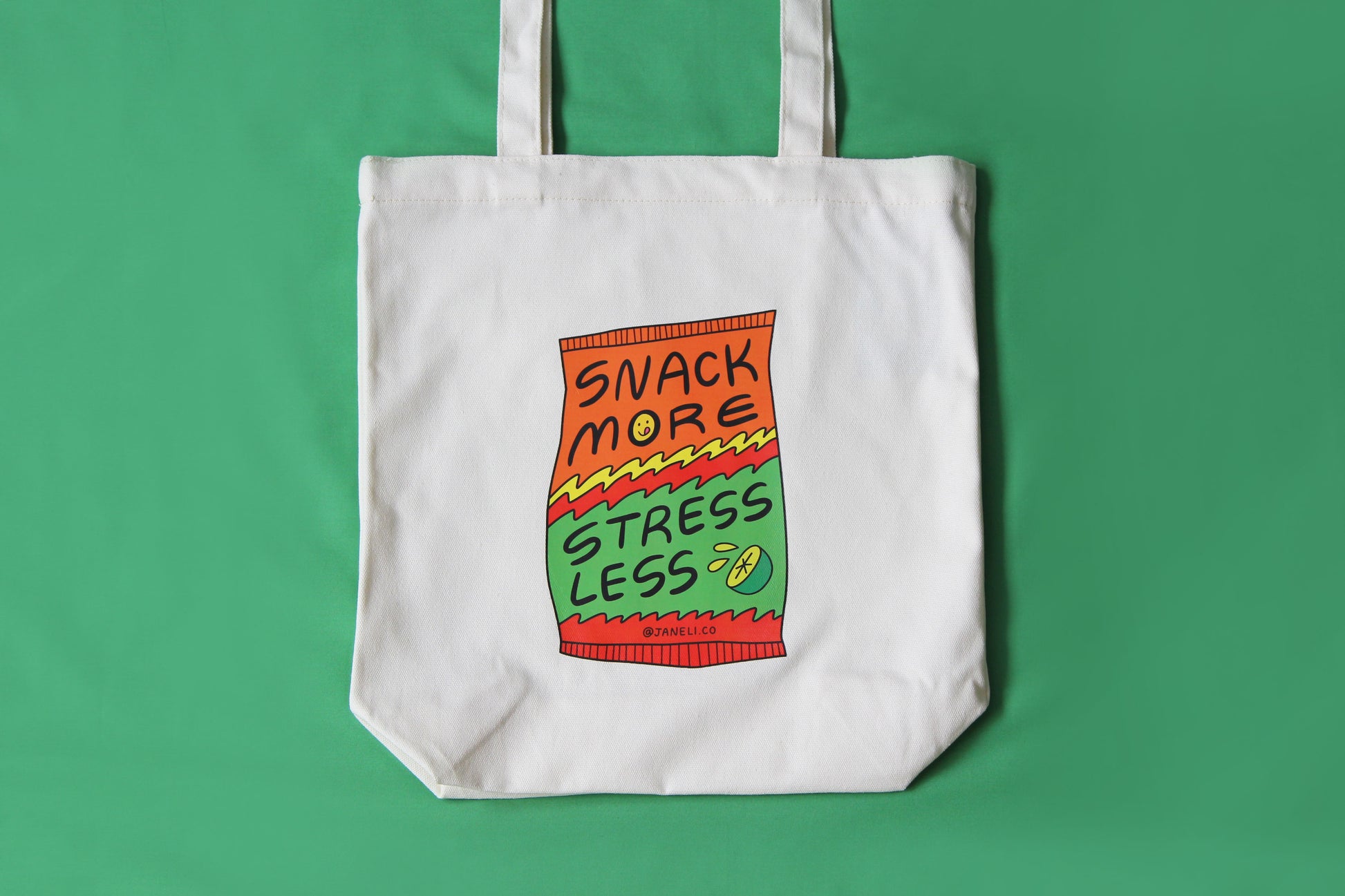 A photo of close up of a natural canvas tote with a chip bag that says "Snack More Stress Less" on it over a green background.