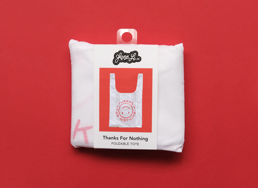 A photo of a white foldable grocery bag with an upside down smiley face that says "Thanks for nothing" packaged in a belly band over a red background.