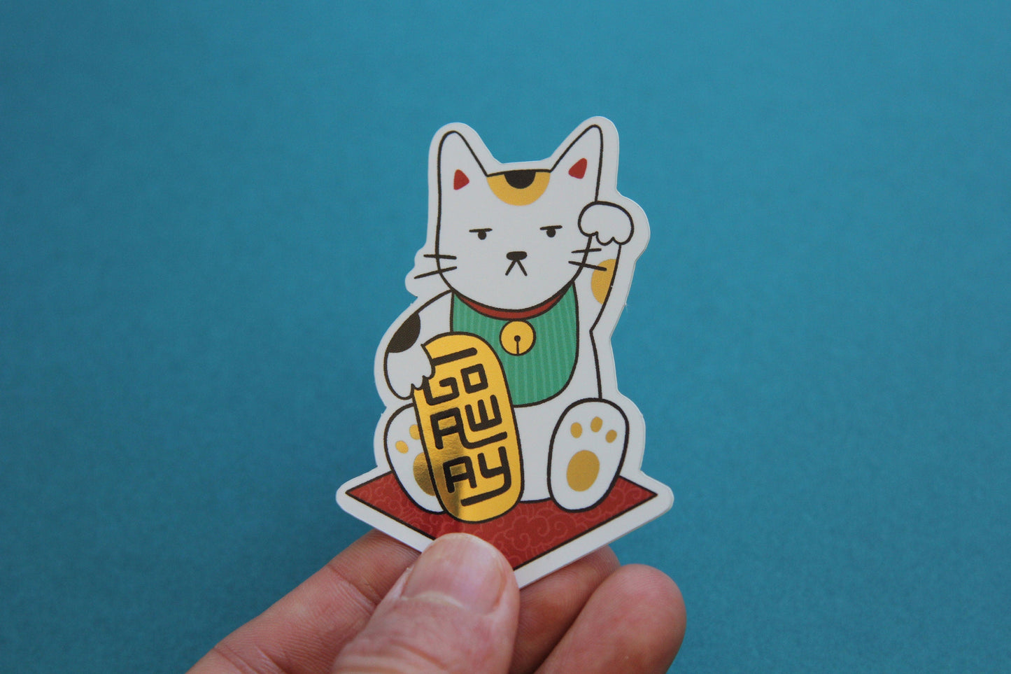 A hand holding a JaneLi.Co sticker that shows a Maneki Neko cat holding a metallic gold bar that says "Go Away" over a teal background. 