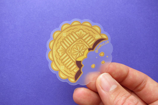 A hand holding a JaneLi.Co sticker of a mooncake over a purple background. 