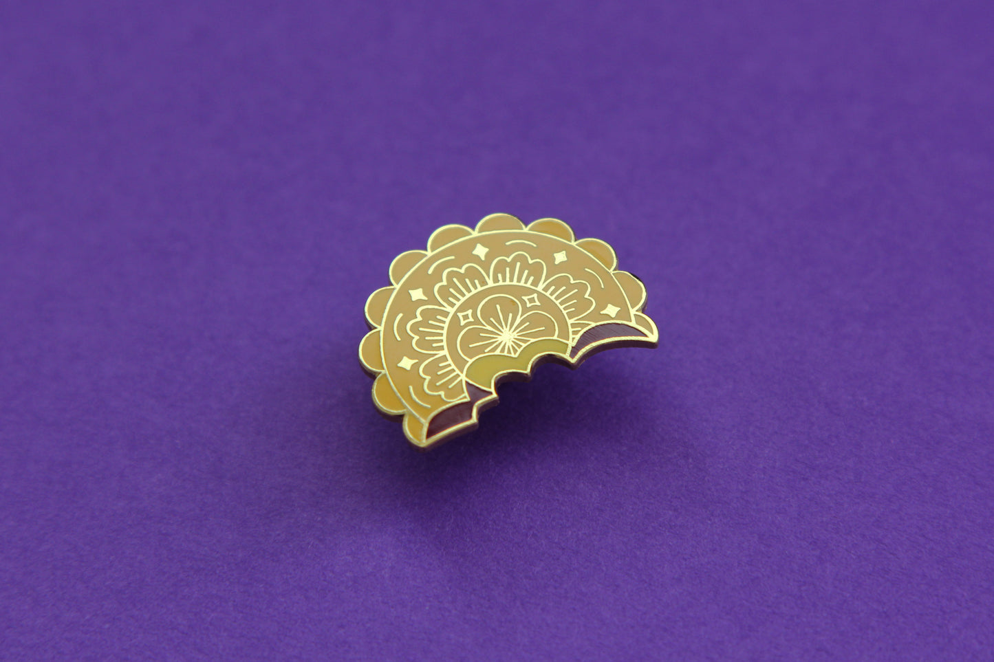 An enamel pin showing a mooncake with a bite out of it over a purple background.