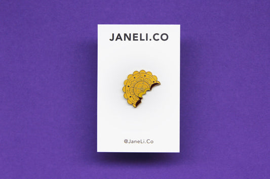 An enamel pin showing a mooncake with a bite out of it on a white JaneLi.Co backing card over a purple background.