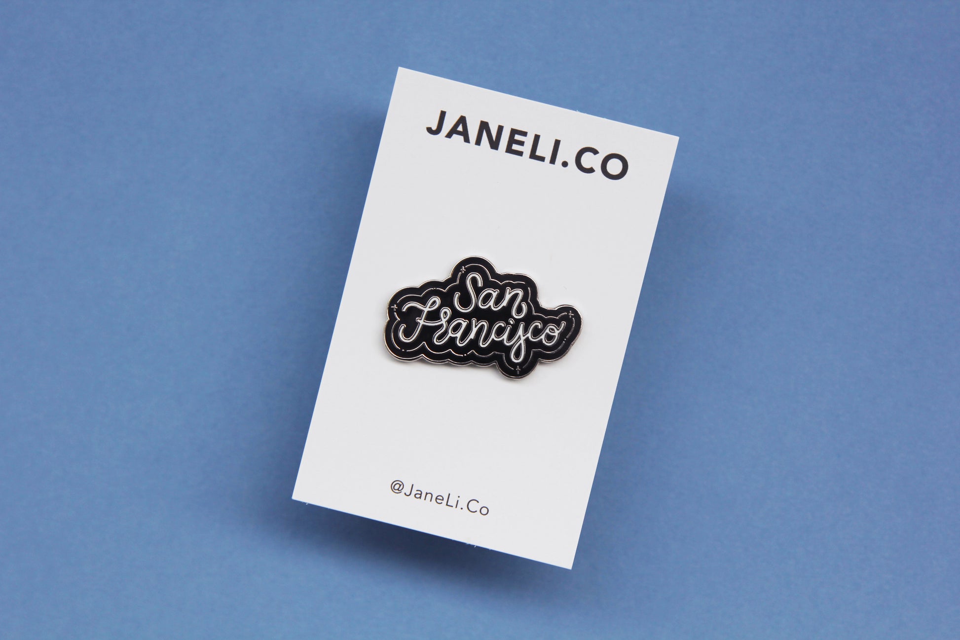 A black, white, and silver enamel pin that says "San Francisco" on a white JaneLi.Co backing card over a blue background.