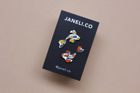 A set of 2 koi fish enamel pins circling on a black JaneLi.Co backing card over a tan background. One koi fish is gold and grey, and the other is scarlet and grey.