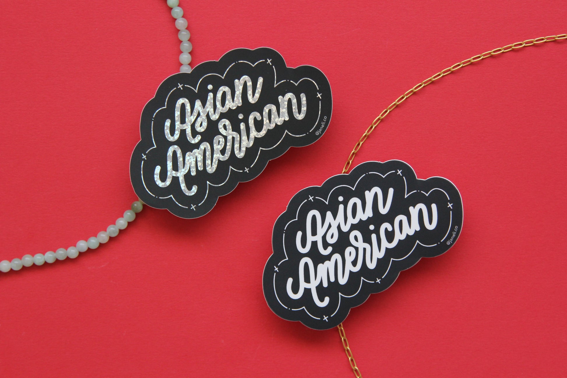 Two JaneLi.Co stickers that say "Asian American" in cursive lettering over a red background with a string of green jade and a thin gold chain. One sticker is black and white, and the other sticker is black and metallic glitter.