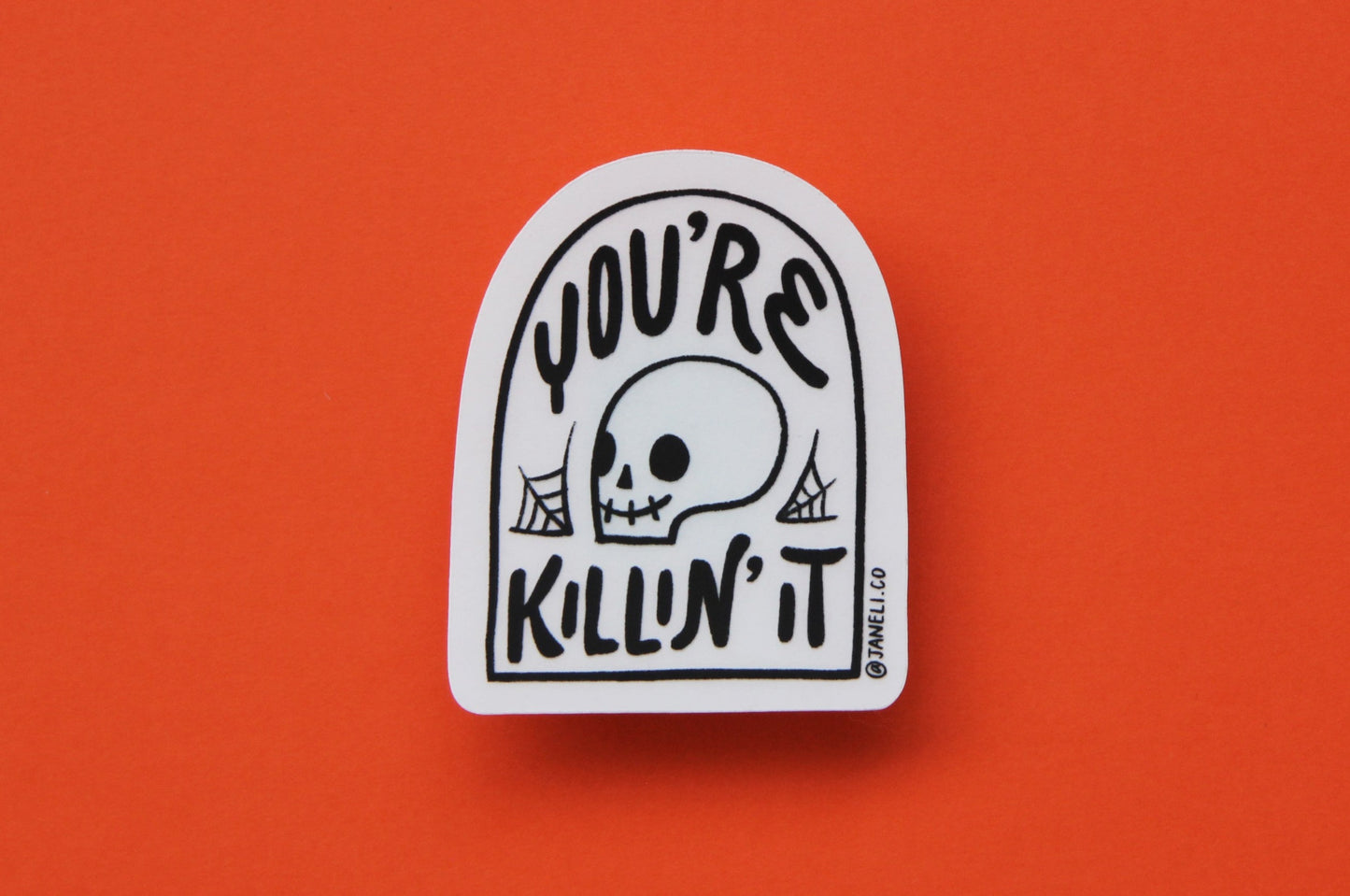 A JaneLi.Co sticker that says "You're Killin' It" in the shape of a tombstone with a skull in it over an orange background.