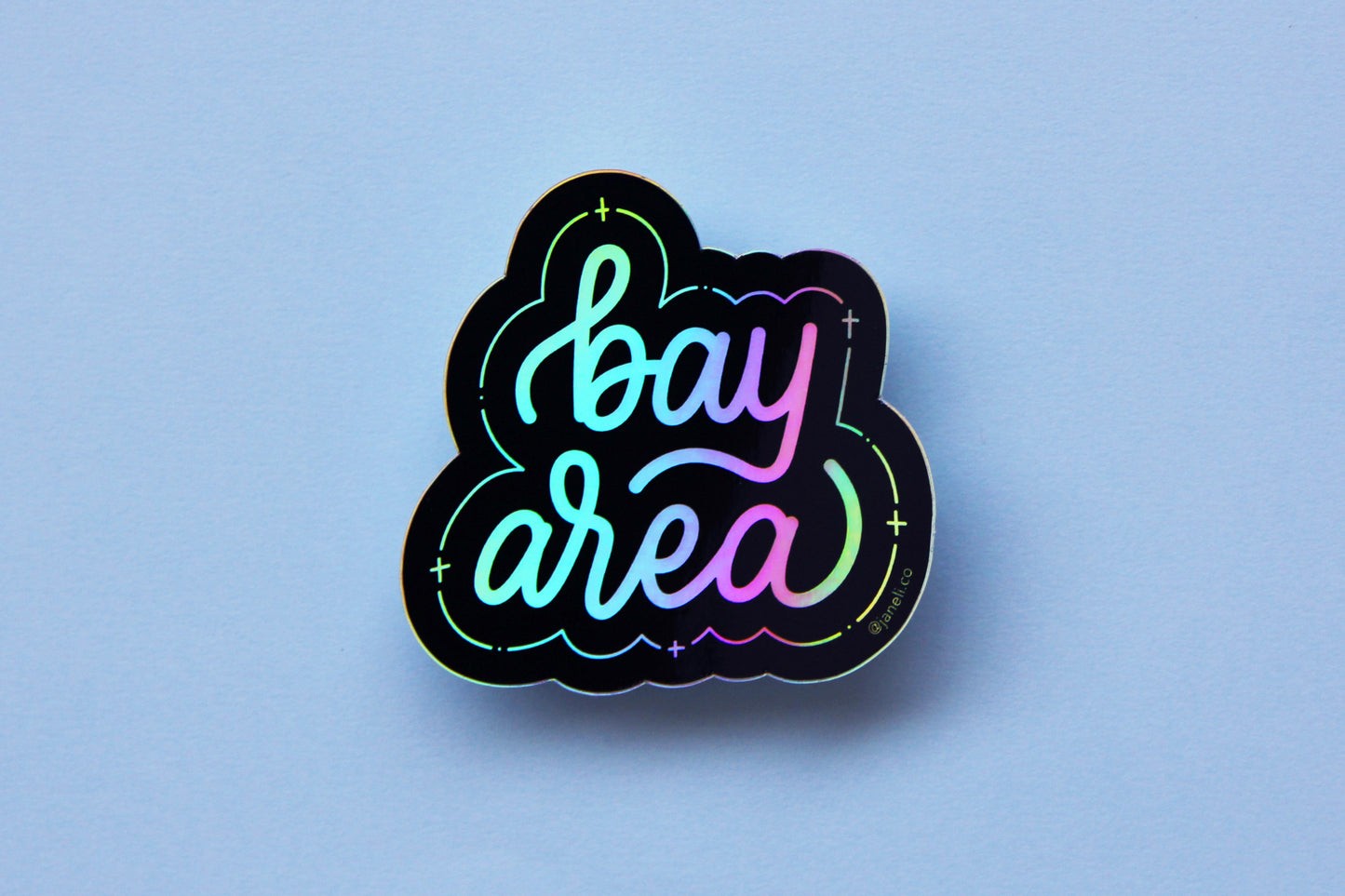 A holographic JaneLi.Co sticker that says "Bay Area" in cursive lettering over a sky blue background. 