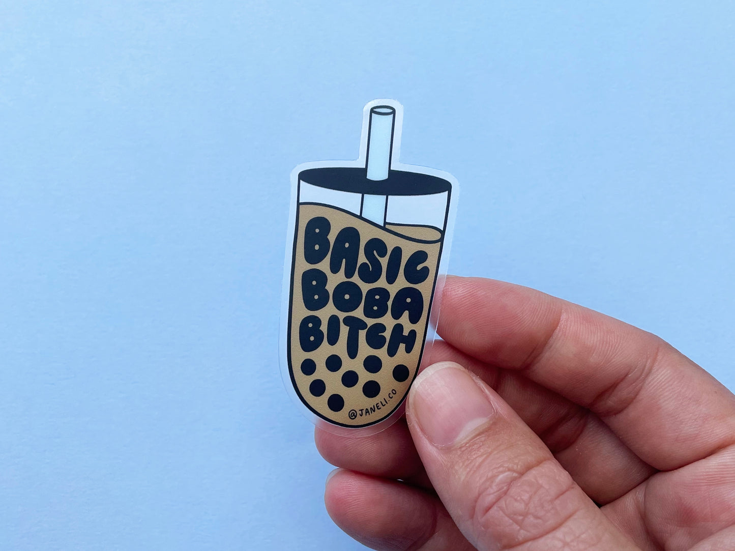 A hand holding a clear JaneLi.Co sticker that says "Basic Boba Bitch" in the shape of a cup of boba over a sky blue background. 